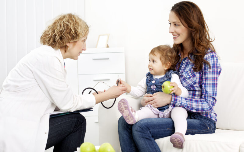 Female doctor pediatrician talking to mother after scheduled baby exam, mother holding baby while baby eating fresh green apple